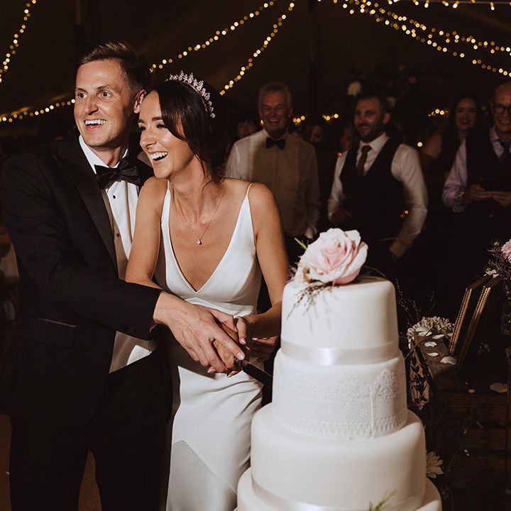 Four tier white iced wedding cake with white ribbon decor being cut by the bride and groom 
