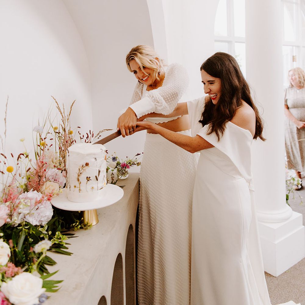 The two brides cut their single tier white iced rustic wedding cake with pressed wildflowers 