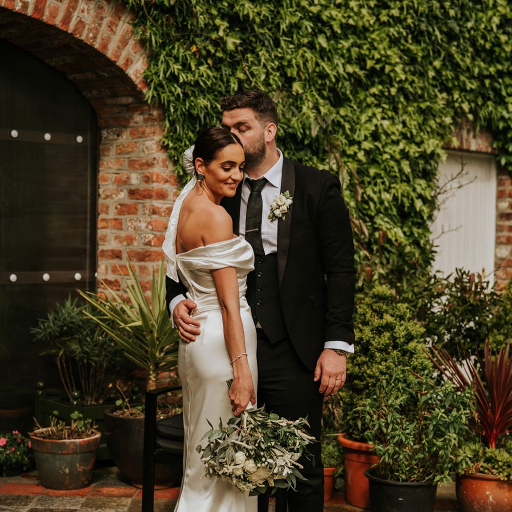 The bride in a classic white dress stands with groom in black tuxedo for their couple portrait together 