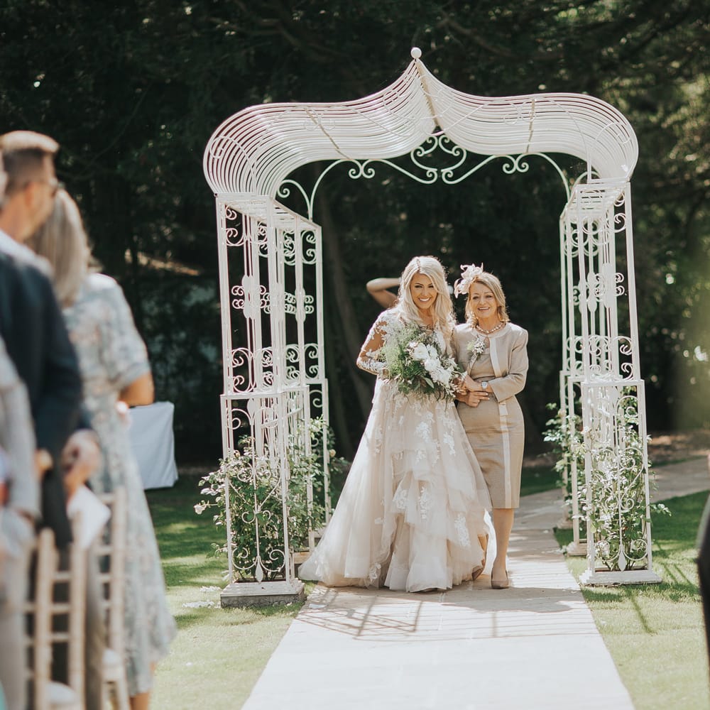 A bride wearing a long tulle layered wedding dress walks down an outdoor aisle with her mom.