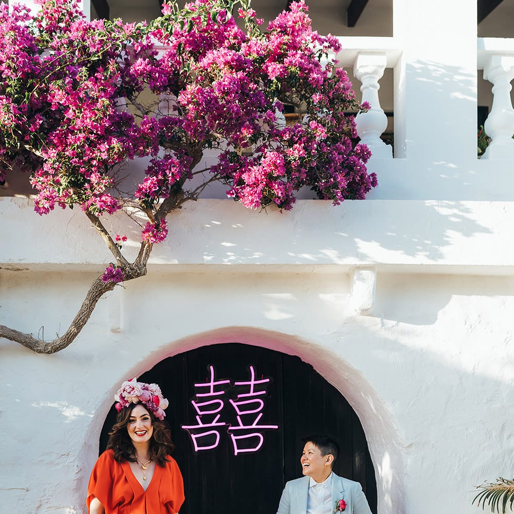 Brides stand in front of pink LED light with Chinese symbols as brides wear red gown and pale blue suit