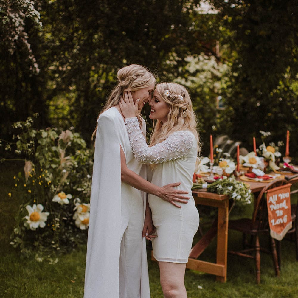Two brides embracing in front of their outdoor tablescape with bold colours and wildflower decor 