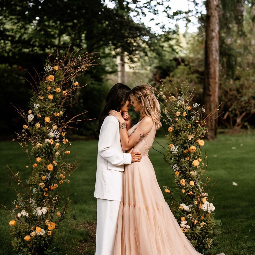 Two brides kissing in between vertical green foliage and yellow flower arrangements at their outdoor wedding ceremony 