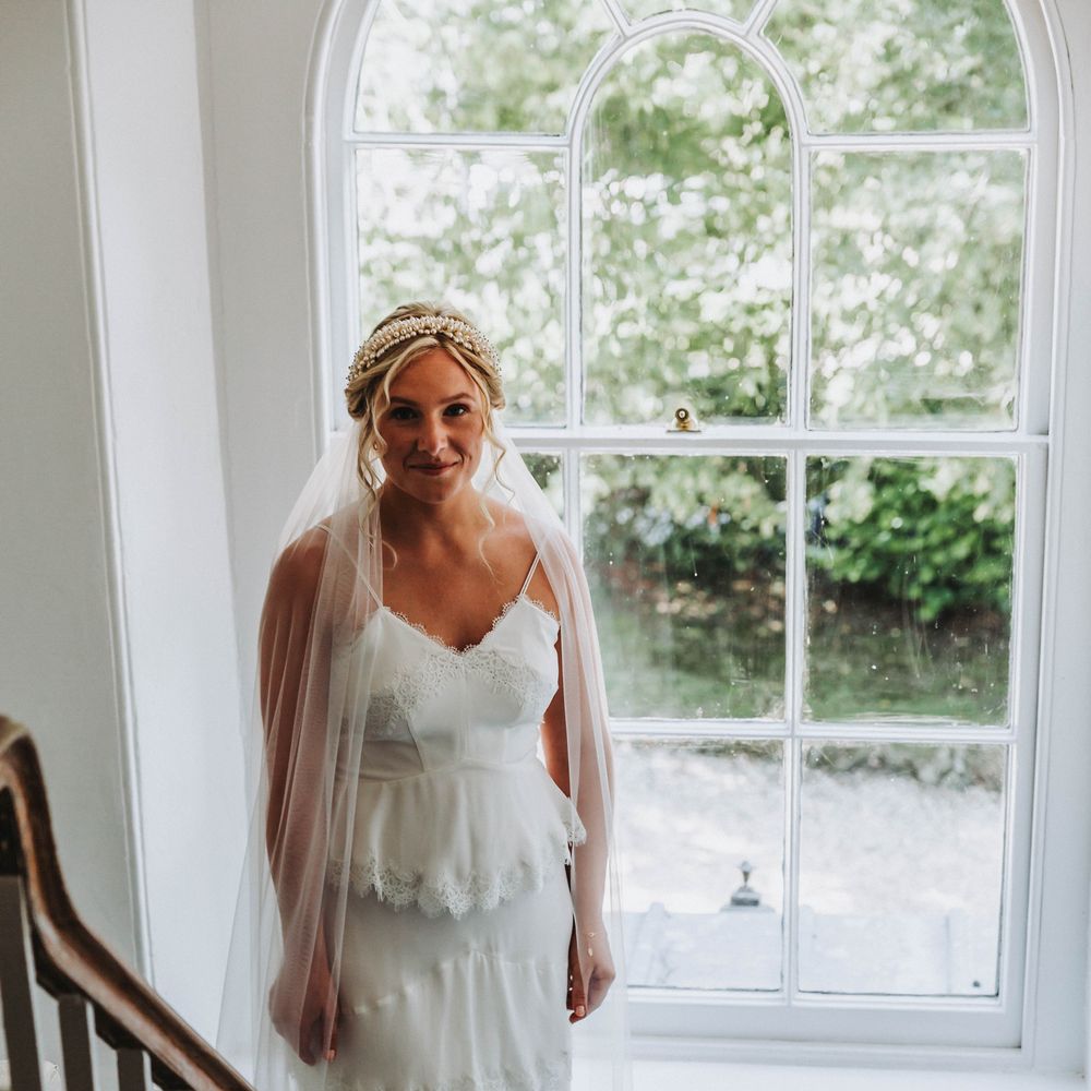 Bride stands in front of window wearing Whistles wedding gown complete with beaded headband on her wedding day