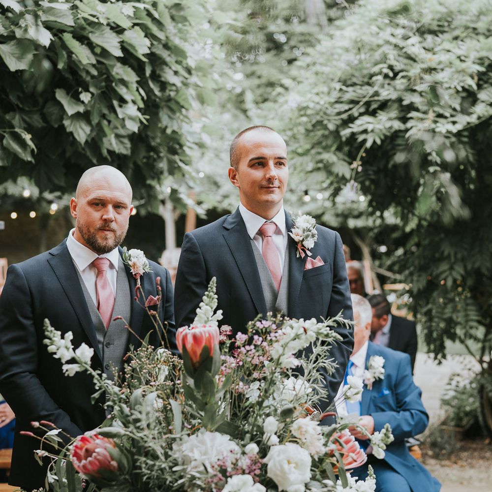 Groom in dark three piece suit with grey waistcoat and pink tie stands with best man by floral bouquet with proteas waiting for bride at Tythe Barn wedding with barn wedding flowers