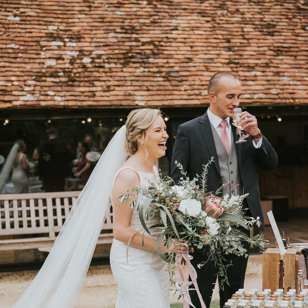 Bride in lace Justin Alexander wedding dress with veil holding pink, white and green mixed bridal bouquet stands next to groom sipping flute of sparkling wine at Tythe Barn wedding with barn wedding flowers