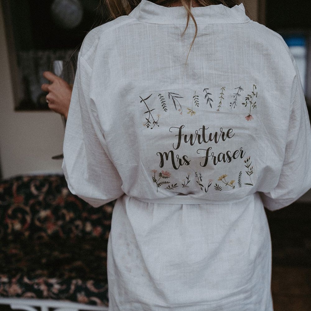 Bride in white dressing gown with writing on the back reading 'Future Mrs Fraser' surrounded by leaf illustrations before Isle of Wight wedding with macrame wedding decor