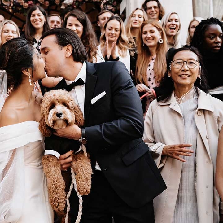 Bride and Groom kiss as they hold dog as the wedding guests cheer and throw hands in the air to celebrate 