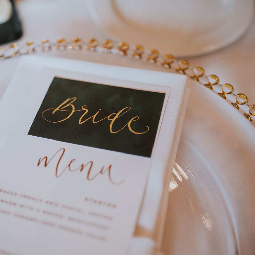Green table place name with gold calligraphy spelling 'Bride' with Menu on white and gold plate for Notley Abbey wedding
