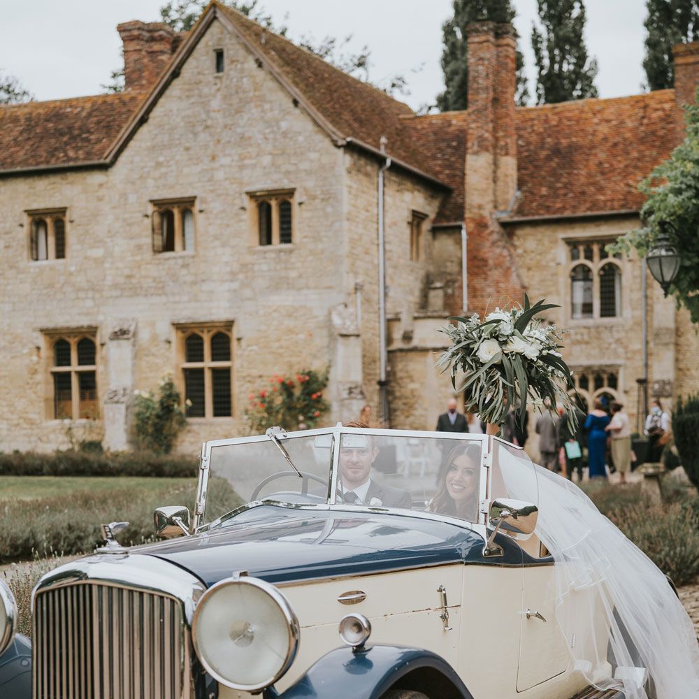 Bride in Pronovias wedding dress, Jimmy Choo wedding shoes and veil holds up white and green bridal bouquet as she sits in vintage car with groom at Notley Abbey wedding