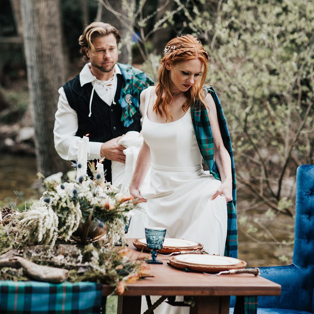Groom dresses in traditional highland wear helping his bride in a taffeta wedding dress sit down at their sweetheart table 