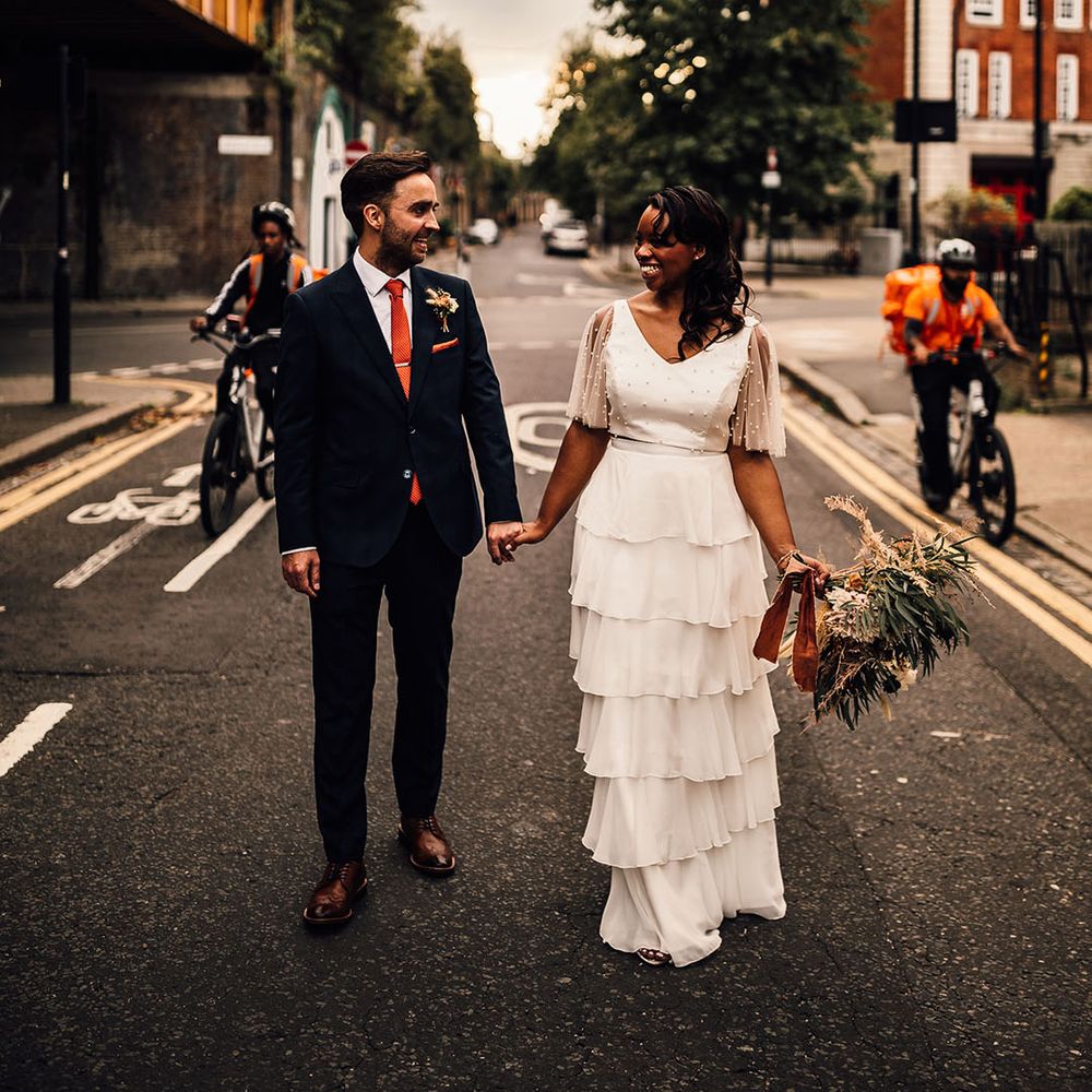 Black bride in a layered skirt and pearl top holding hands with her groom in a navy suit on the streets of London during golden hour 