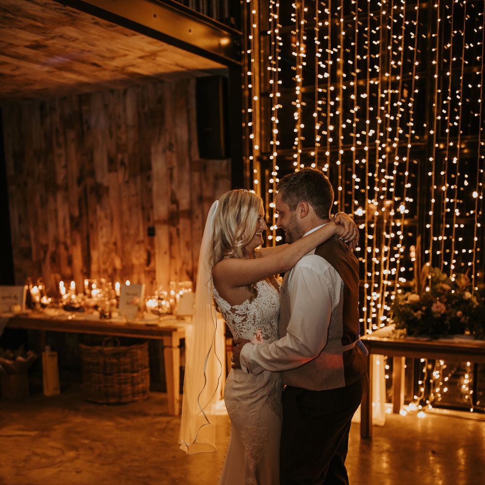 Bride in lace wedding dress with train and veil stands with groom in white shirt and waistcoat in front of fairy light curtain at rustic wedding
