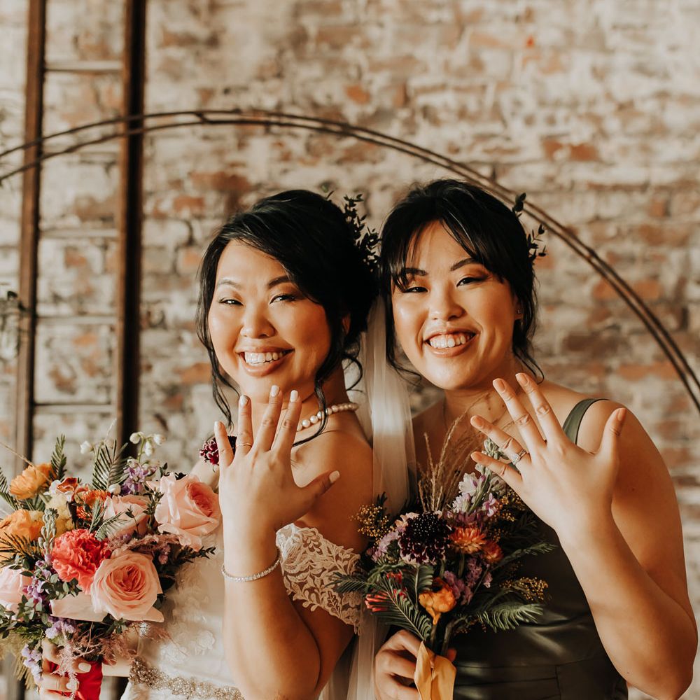 Bride in lace bardot wedding dress holding mixed floral bouquet stands with bridesmaid in sage green satin bridesmaid dress both showing off their rings