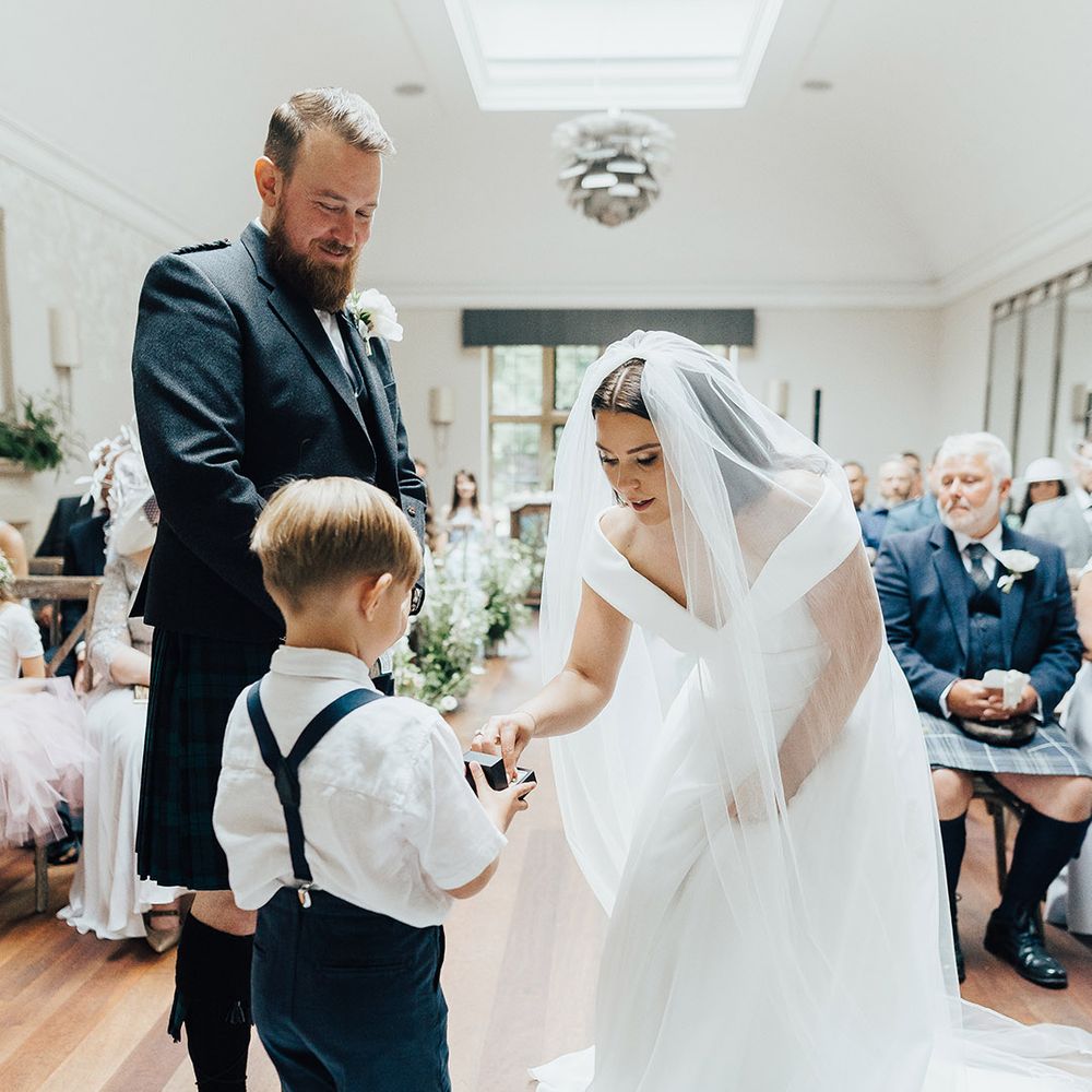 Bride in an off the shoulder Suzanne Neville wedding dress and veil taking her ring out the box from her son ring bearer in navy shorts and braces 