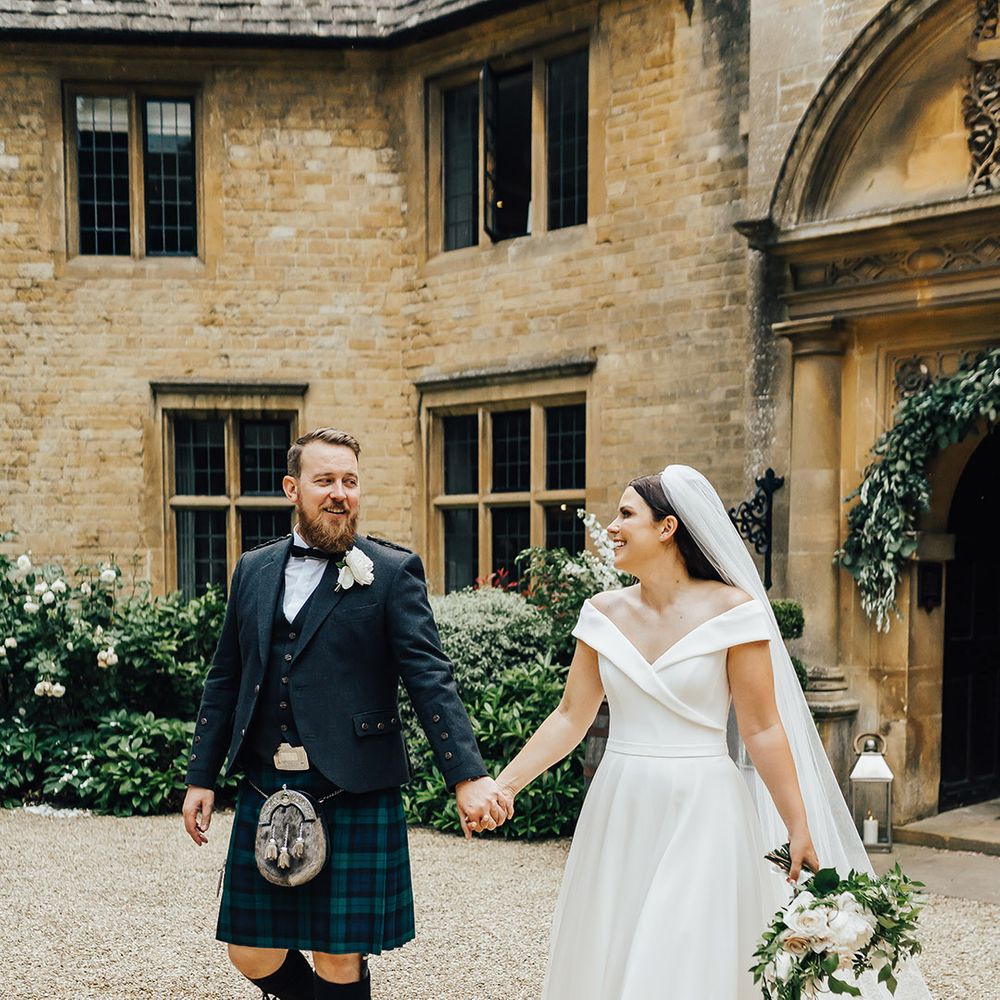 Groom in a green and blue tartan kilt holding hands with his bride in an off the shoulder wedding dress with long veil 