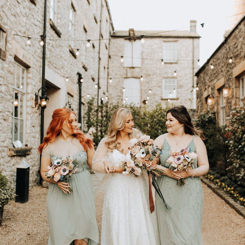 Bride in v neck wedding dress encrusted with pearls walks with bridesmaids in sage green bridesmaid dresses all holding mixed pastel wedding bouquets