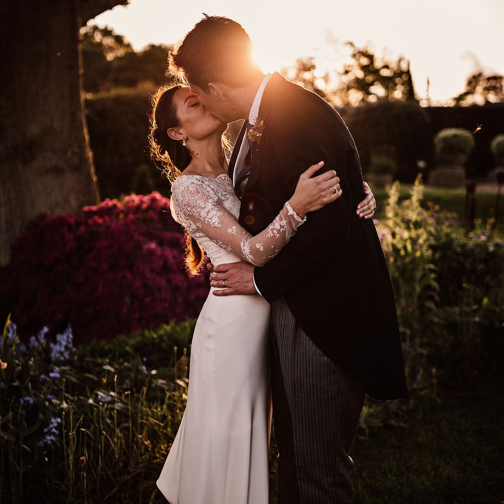 Bride and groom share a kiss at golden hour with groom in morning suit and bride in sheer lace long sleeve wedding dress 