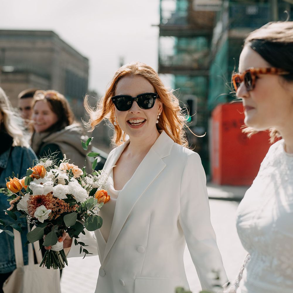 Bride looks toward camera wearing sunglasses whilst holding bright floral bouquet 