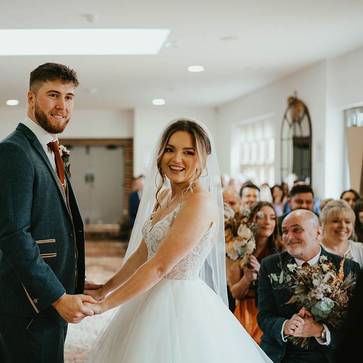 Bride and groom smile happily as they hold hands for their ceremony as both of their fathers smile at each other 