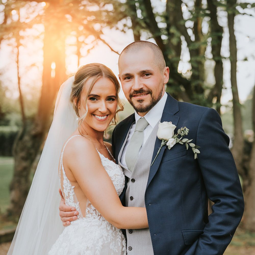 Bride in a lace wedding dress with the groom in a dark blue suit and white rose buttonhole with a light grey suit and green tie 