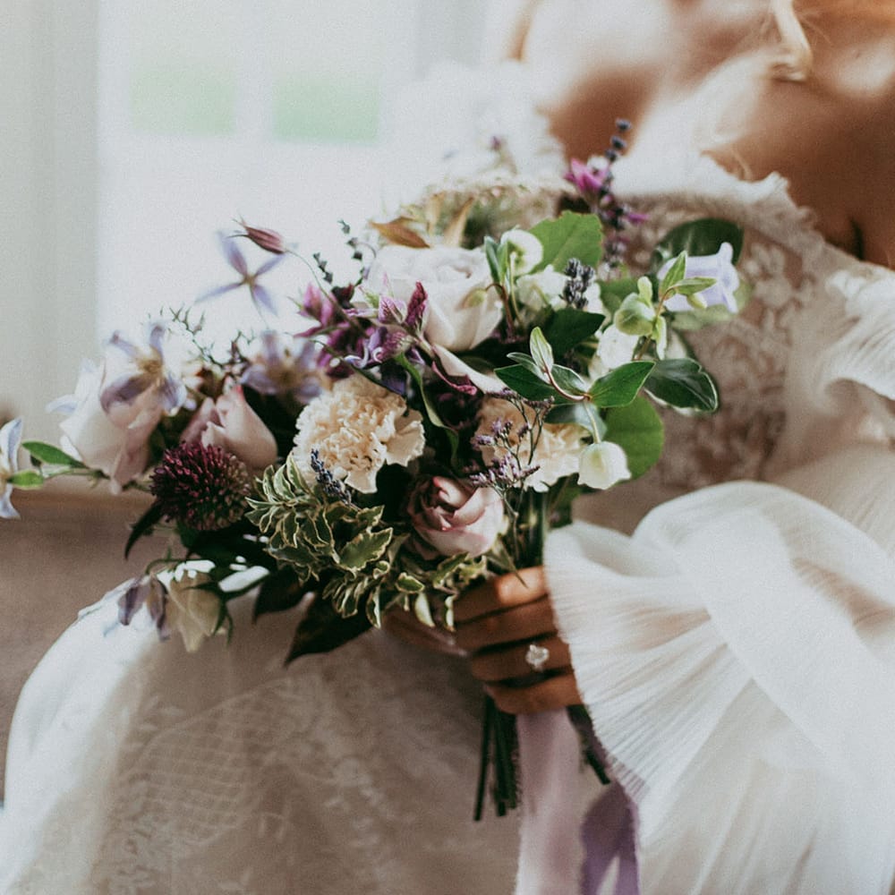 Bride holding mixed neutral floral bouquet with white carnations, purple tulips, roses and foliage 