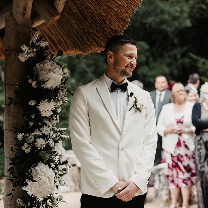 The groom in a white suit jacket and black tie with a white rose buttonhole stands smiling as he sees the bride walking to him 