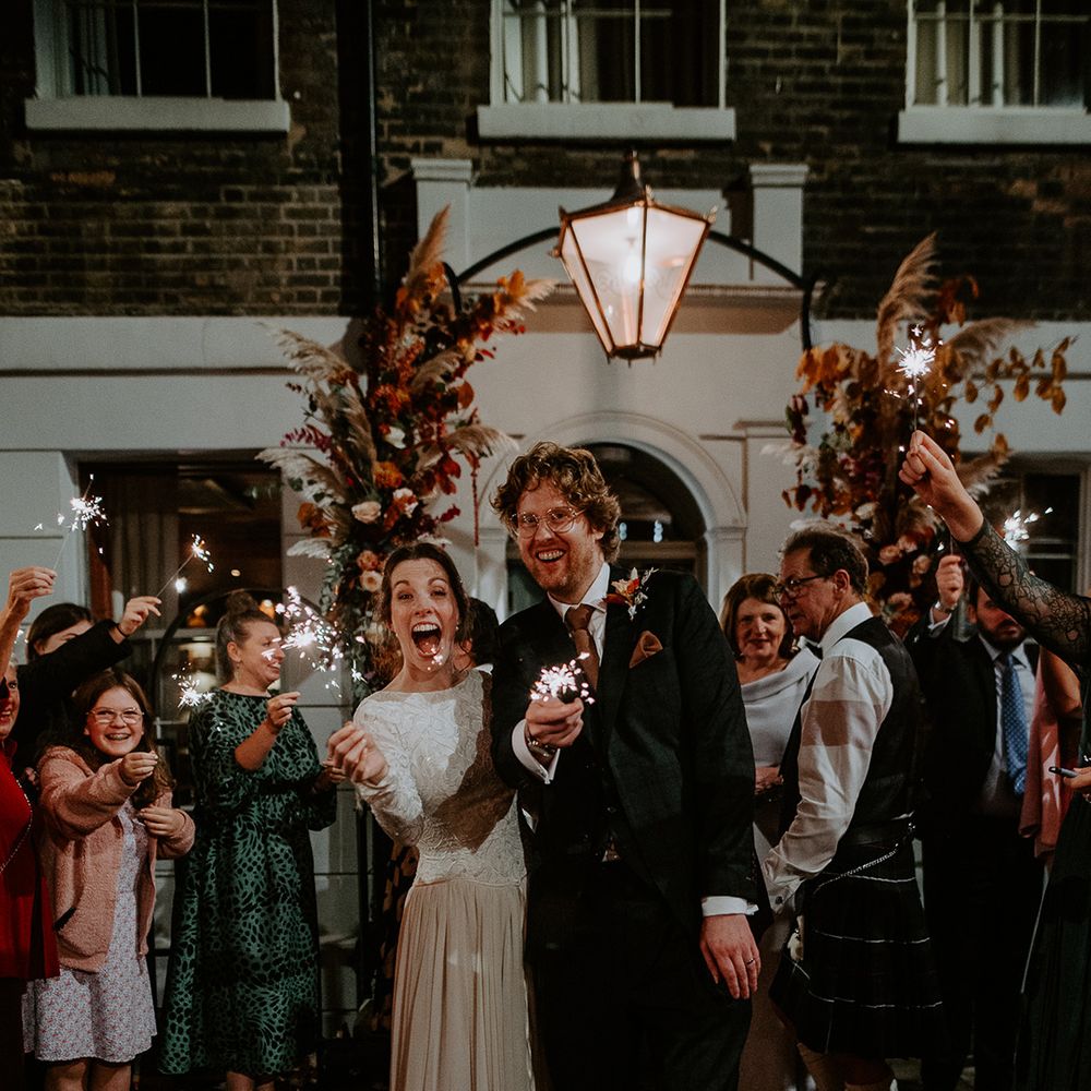 Bride in long sleeve lace vintage wedding dress and closed toe satin heels and groom in dark suit with orange tie and autumnal boutonniere doing sparkler exit from autumnal intimate London wedding