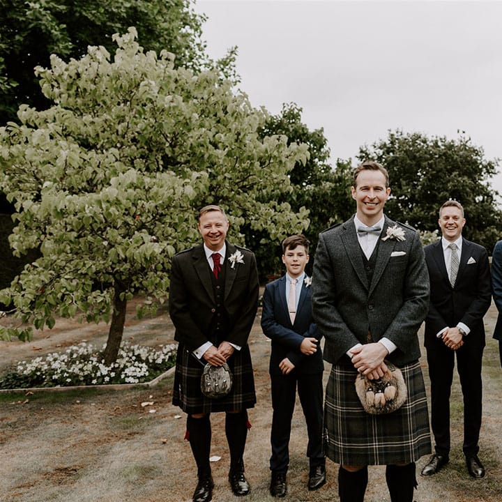 Groom in tweed suit jacket and pale blue bow tie and kilt stand smiling in front of groomsmen and family members 