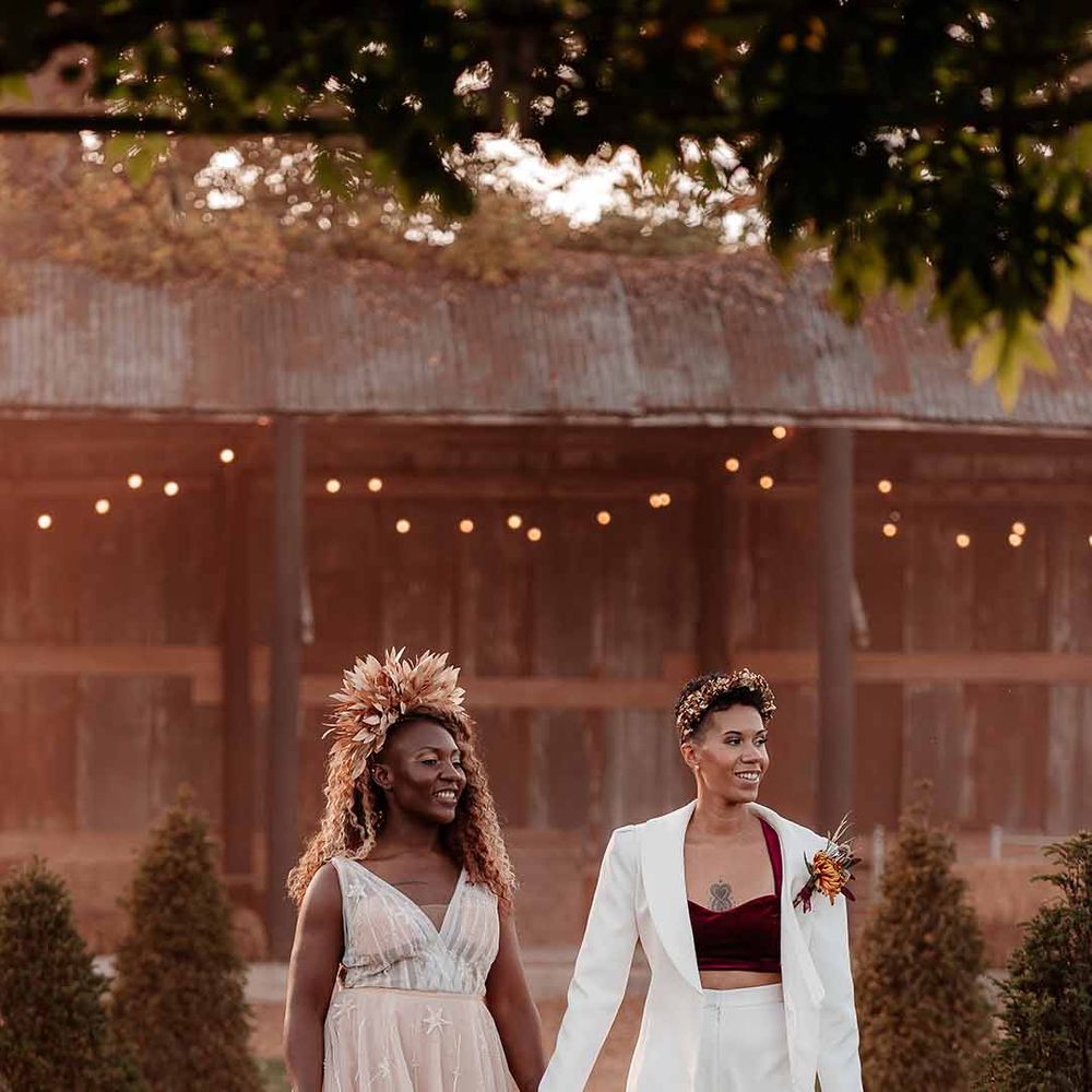Bride in elegant white silk suit and deep burgundy velvet crop top with autumnal dried floral boutonniere holding hands with bride in sleeveless floaty peach gown with embroidered stairs and gold leaf crown  