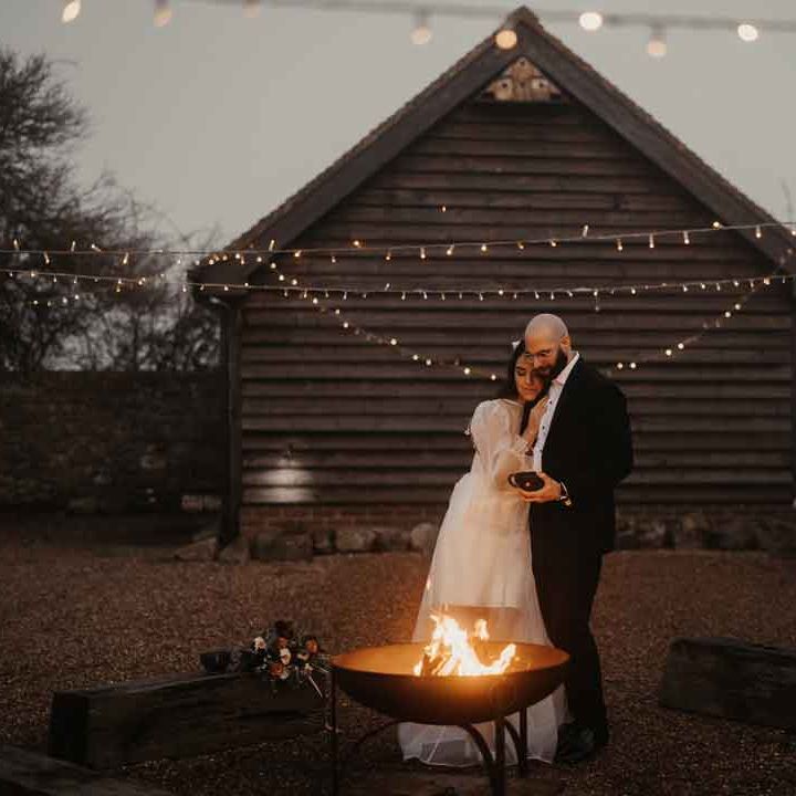 Bride in square neck wedding dress with sheer puff sleeves embracing groom in in classic black tuxedo with black bowtie standing by the fire pit 