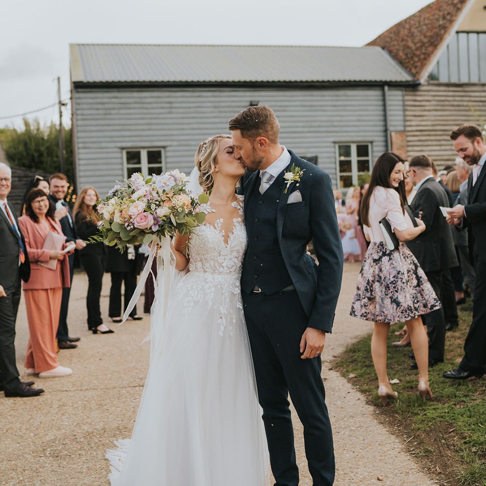 The bride and groom share a kiss after their confetti exit with the bride in an illusion lace wedding dress and navy suit 