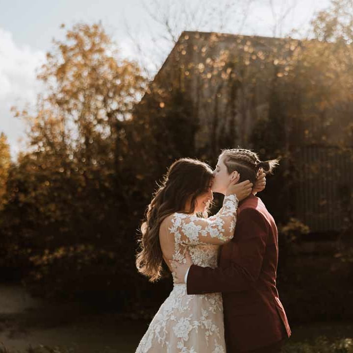 Bride wearing maroon bridal suit with grey waistcoat, dusky pink tie and autumnal dried flower boutonniere kissing bride on the forehead wearing long sleeve lace illusion sleeve wedding dress with overlay 
