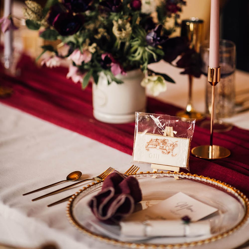 Iced wedding biscuits with gold cutlery, gold charger plate, burgundy napkin and table runner with purple floral arrangement and taper candle
