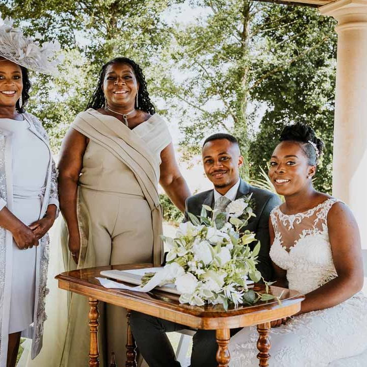 Bride in lace and mesh boatneck wedding dress with silver bridal hair accessories sitting with groom in charcoal grey grooms suit, light grey patterned waistcoat, light grey patterned tie and large white rose and foliage boutonniere and mothers of the bride and groom signing the register at Tredudwell Manor