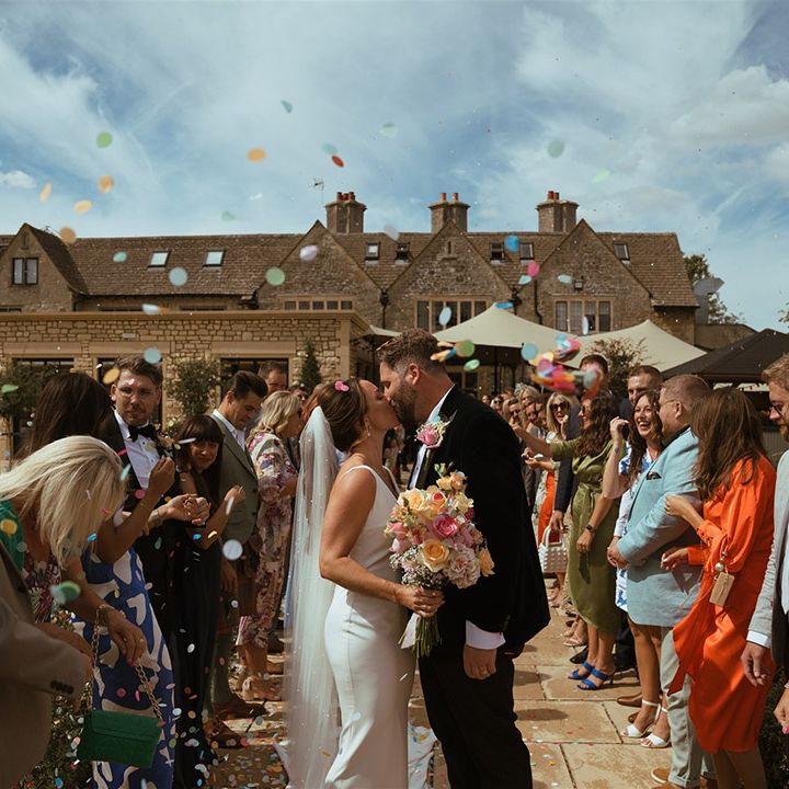 The bride and groom kiss at their confetti exit from their outdoor ceremony with guests throwing confetti 