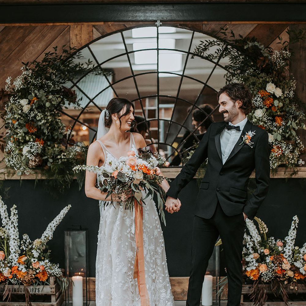 Bride in floral lace wedding dress standing with groom in a black tuxedo at Ghyll Barn Lake District wedding venue 