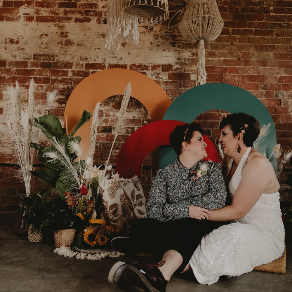 Brides in a jumpsuit and patterned shirt sitting by a colourful altar styled with dried flowers 