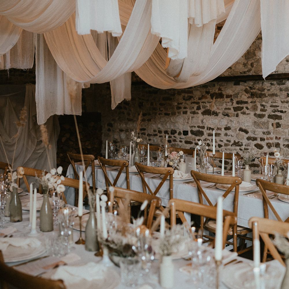 Exposed brick wedding venue, with wooden chairs, fabric ceiling, neutral flowers and taper candles 