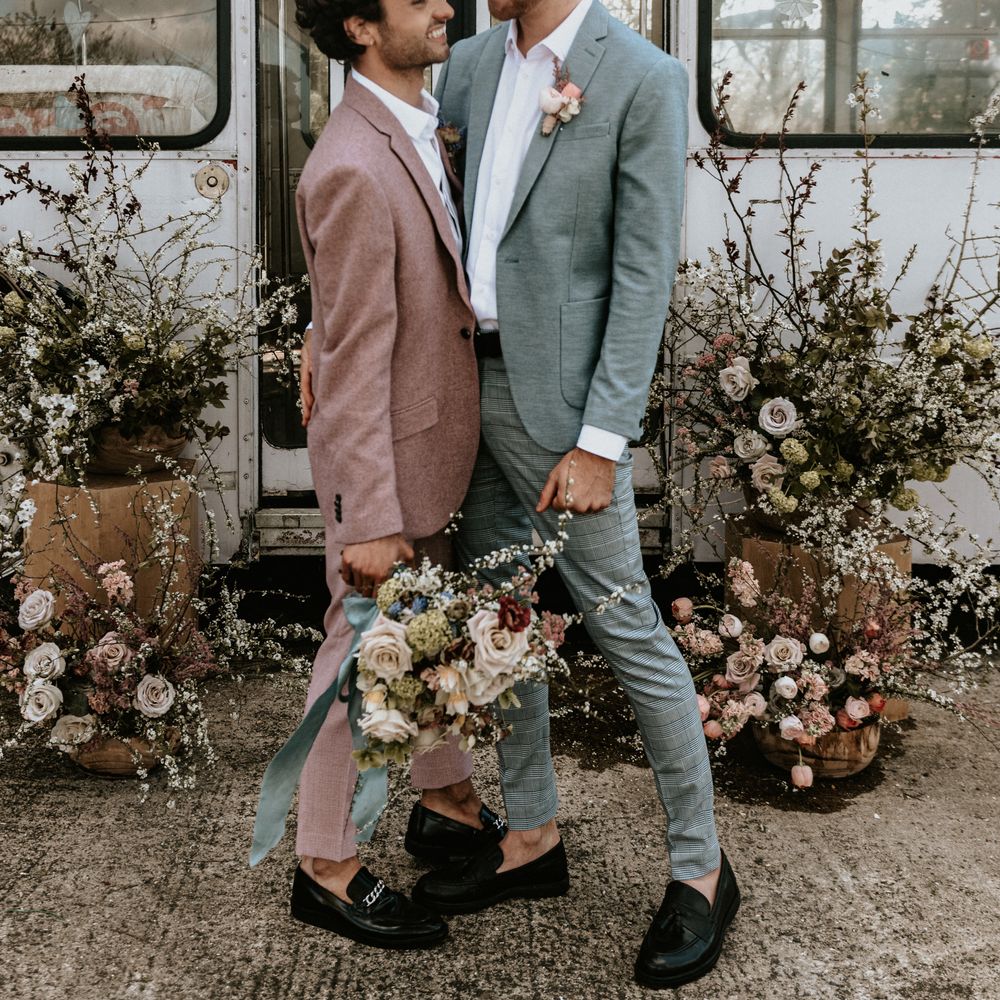 Grooms smile and laugh in front of rustic floral installations whilst holding floral bouquet 