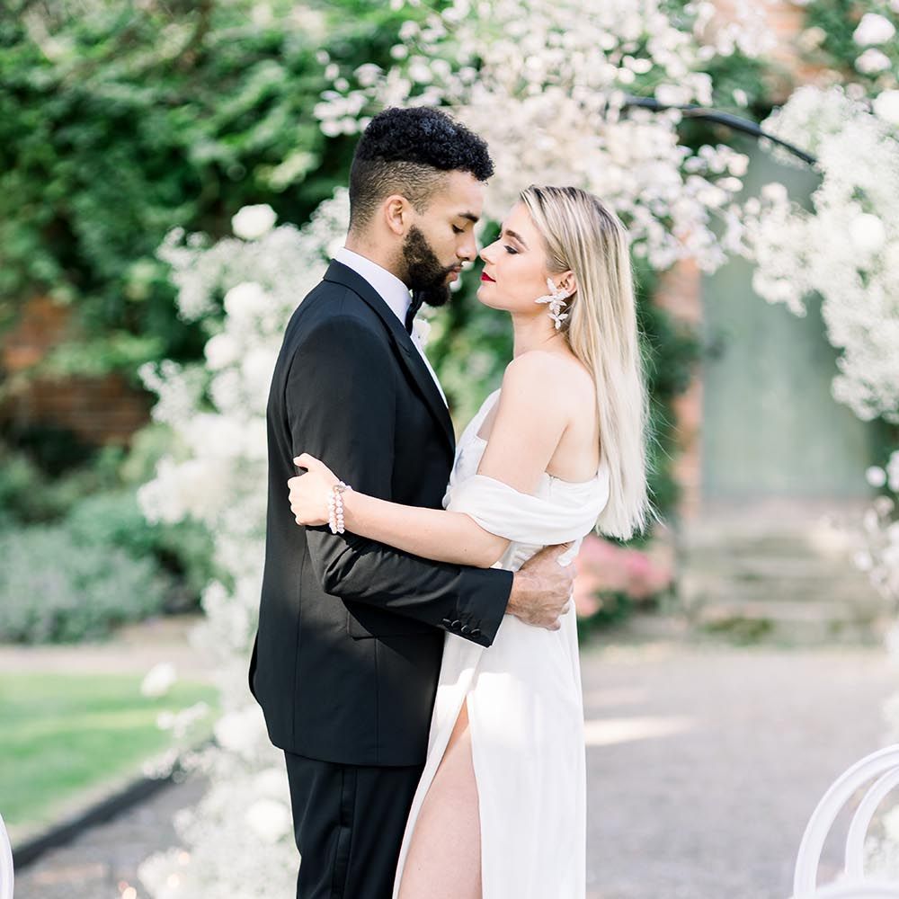 Bride in an off the shoulder wedding dress with front split embracing her husband in a black tie suit