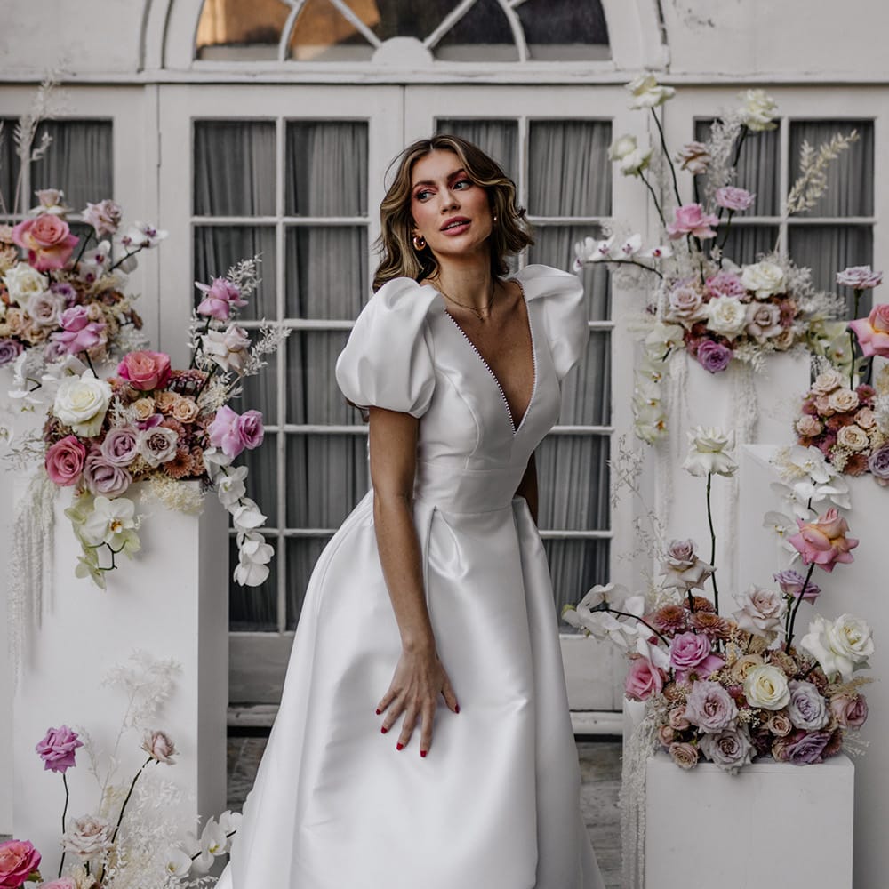 Stylish bride in a Love In Lace bridal gown with puff sleeves and plunging neckline surrounded by pink wedding flowers 