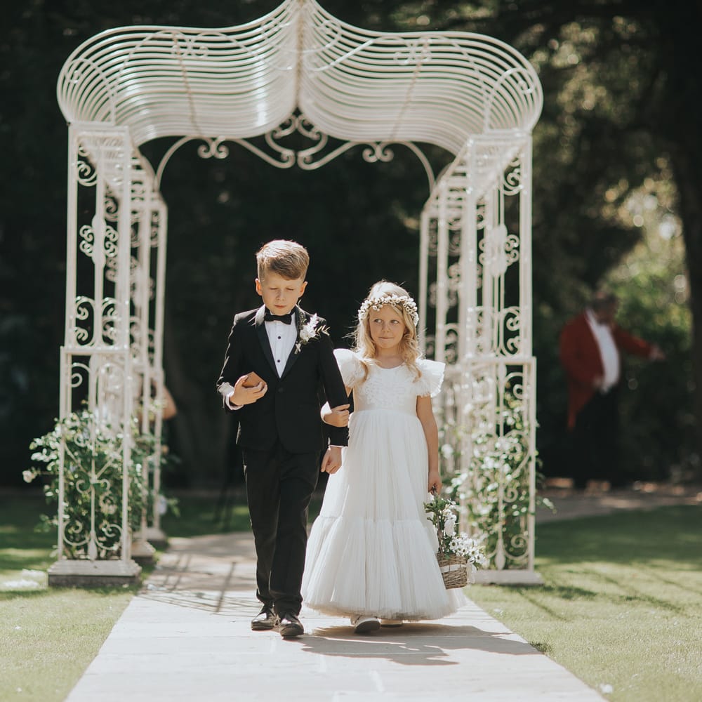 A young girl in a white dress walks down a wedding aisle with a young boy in a black tuxedo. The aisle is outside.