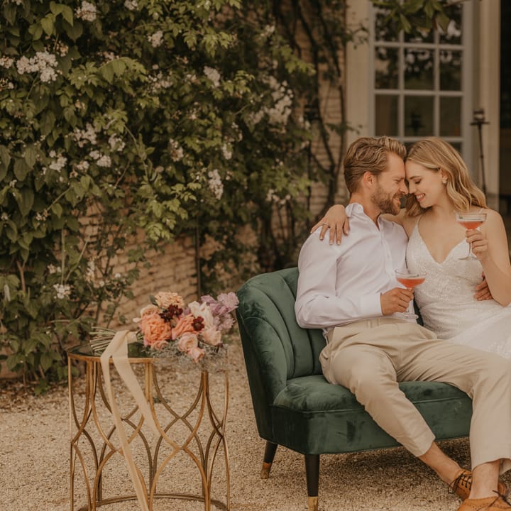 Bride and groom sitting on a green velvet sofa sipping cocktails at their outdoor wedding 