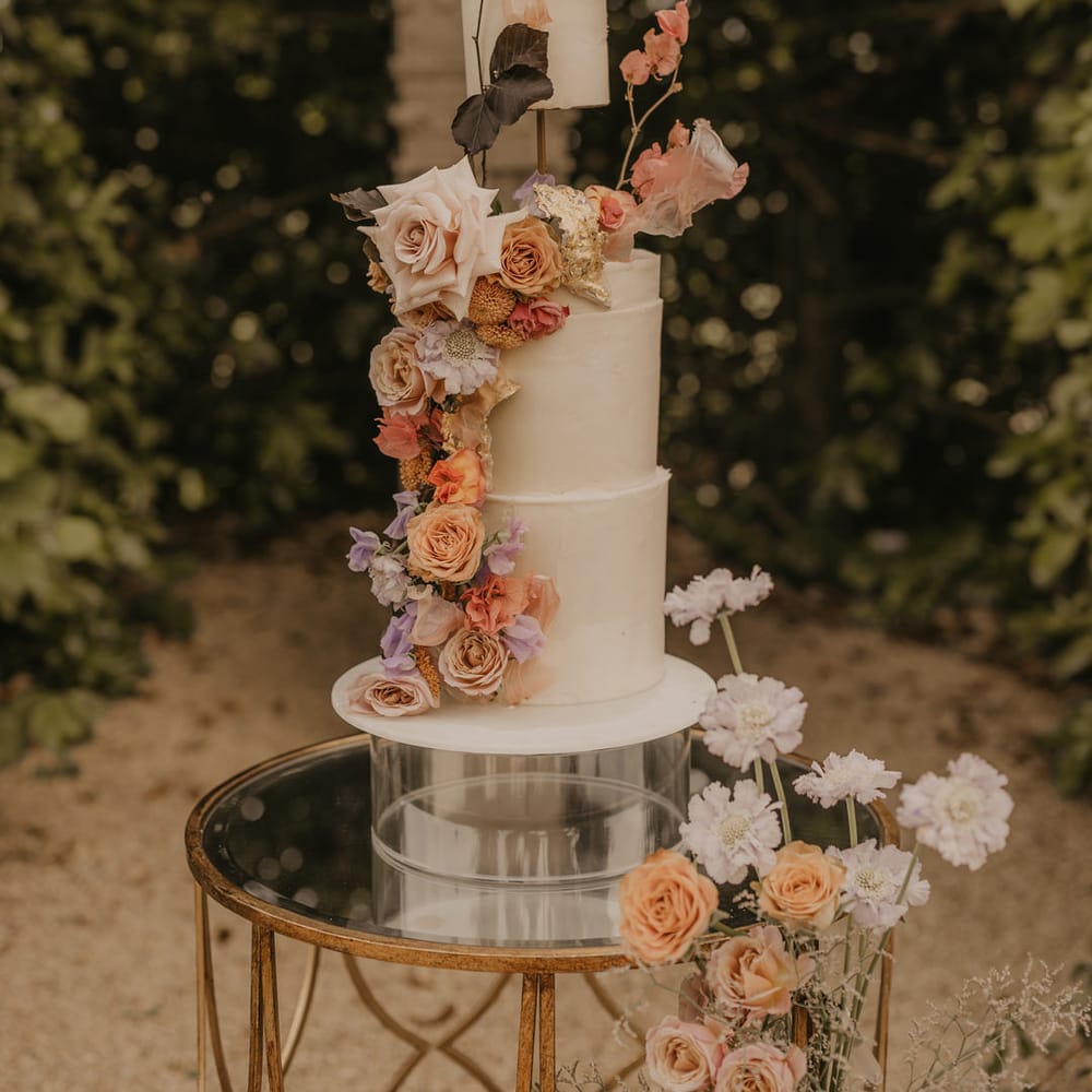 Tall three tier buttercream wedding cake with elevated layer decorated with pastel orange, peach, coral and pink wedding flowers 