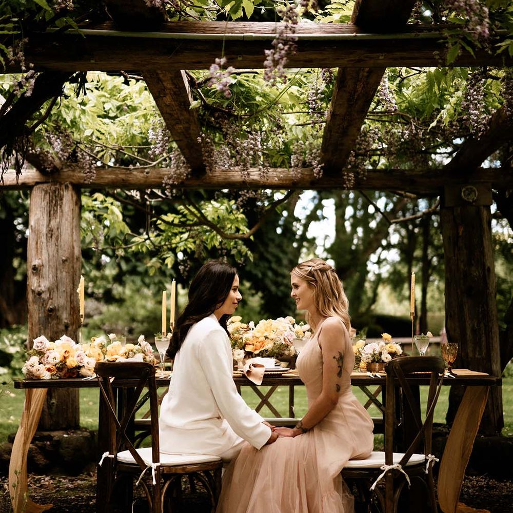 Bride in a white suit and blush pink wedding dress sitting at their al fresco wedding reception table under a pergola 