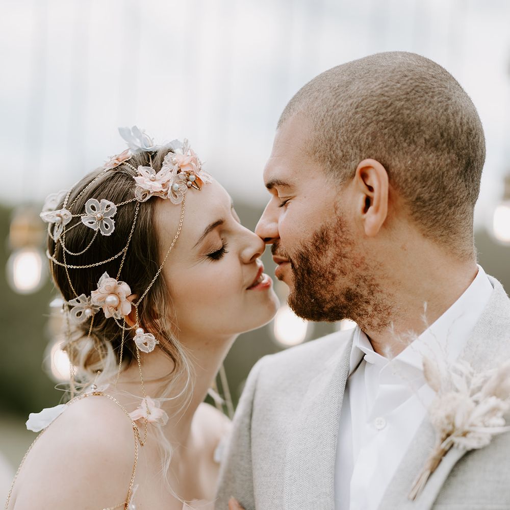 Bride in an intricate chain and flower headdress kissing her husband in a grey suit 