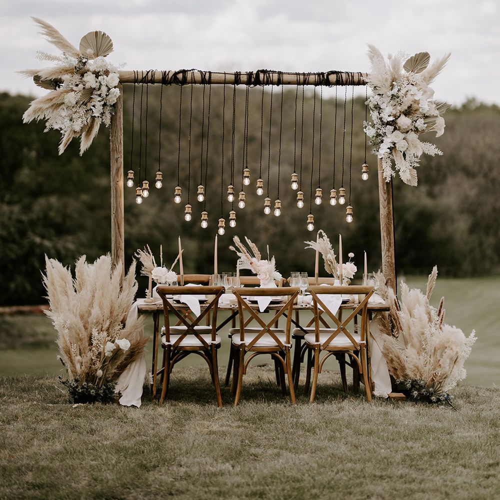Outdoor wedding reception with wooden table and chairs, dried palm leaves and pampas grass floral decor, and Edison bulb light installation 