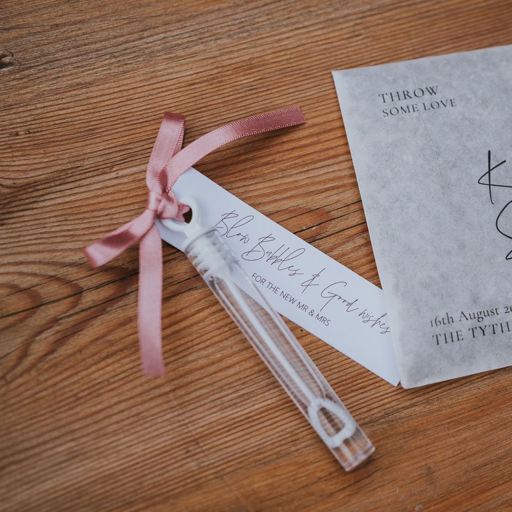 Grey paper personalised confetti bag next to bubbles tied with pink ribbon at Tythe Barn wedding with barn wedding flowers