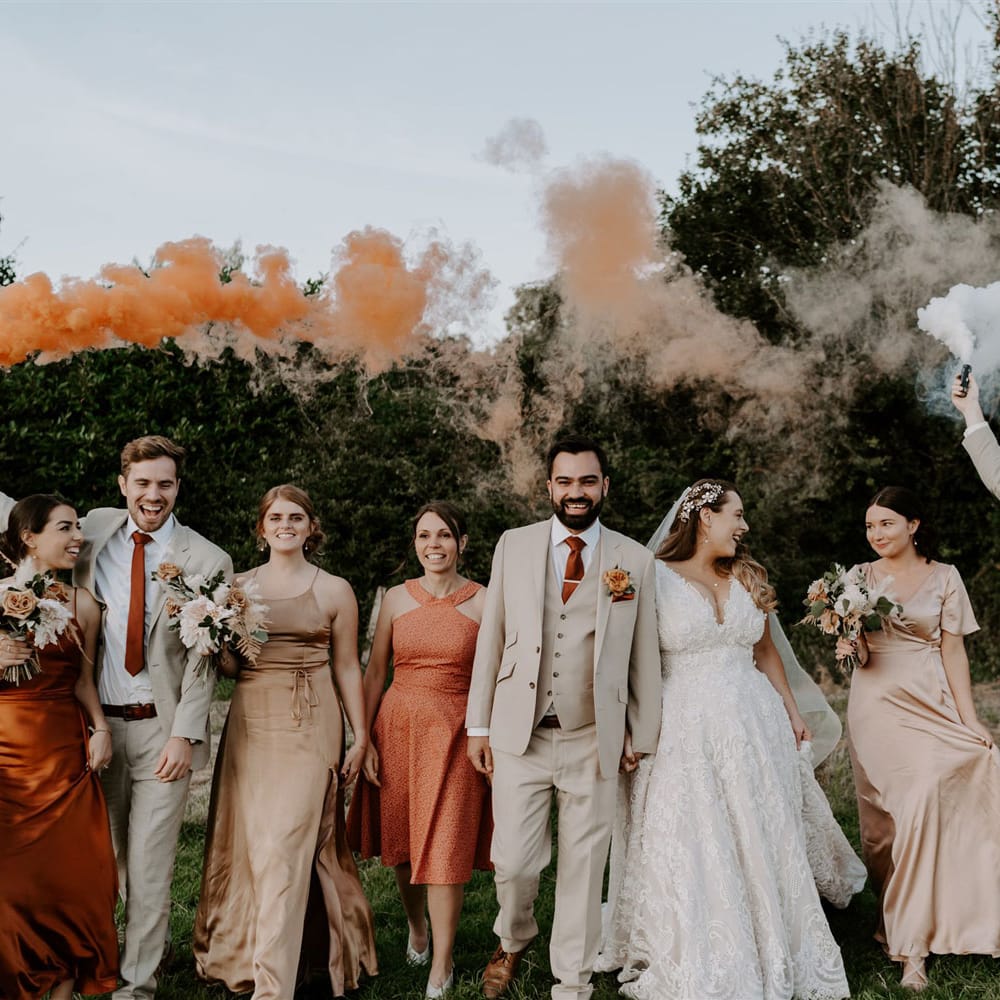 Bride in homemade lace wedding dress holds hands with groom in beige three piece suit as they walk with groomsmen and bridesmaids through field whilst holding orange and white smoke bombs